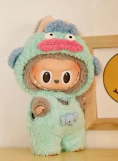 a stuffed animal wearing a green outfit and holding a blue object in front of a wall