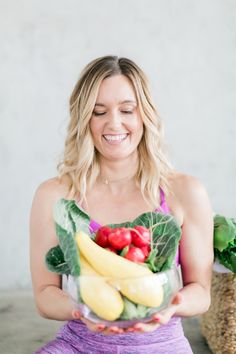 a woman sitting on the floor holding a bowl full of fruit and veggies