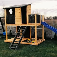 a wooden play house with a slide and climbing frame in the grass next to a brick wall