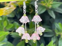 pink flower earrings with green beads hanging from hooks in front of some leaves and bushes