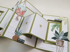 an open book with pictures and plants on the pages, hanging from a hook in front of a white brick wall