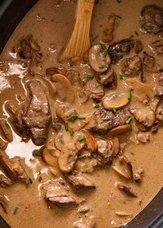 a crock pot filled with meat and mushrooms covered in gravy next to a wooden spoon