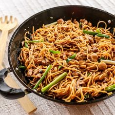 a wok filled with noodles and meat on top of a table next to a wooden spatula