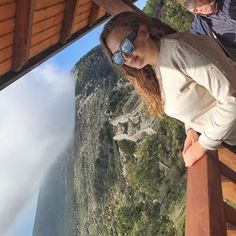 a woman standing on top of a wooden balcony next to a cliff side with mountains in the background