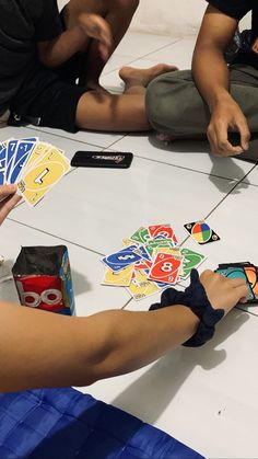 several people sitting on the floor playing cards with their hands and fingers, while one person is holding a cell phone