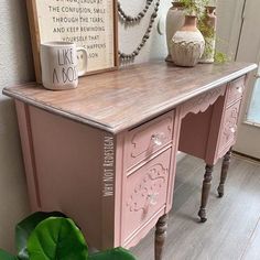 a pink desk with a coffee mug on it next to a potted plant and framed art