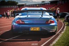 a blue sports car parked on the side of a track with people walking around it