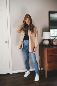 ashley wearing a tan shacket over a black tank, medium wash denim, and white sneakers with a brown beanie standing next to a vintage wood dresser with a white table lamp What To Wear With Shacket, Fall Style 2023 Women Casual, Oatmeal Shacket Outfit, Fall Shaket Outfits, Courdoroy Shacket Outfit, Minimalist Neutral Outfits, High 60 Degree Weather Outfit, Tan Shacket Outfit Women Fall, Neutral Shacket Outfit