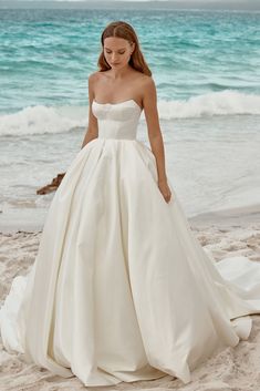 a woman is standing on the beach wearing a wedding dress and looking down at her