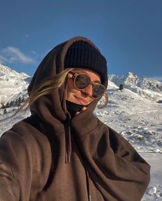 a woman wearing sunglasses and a brown hoodie is standing in front of snow covered mountains