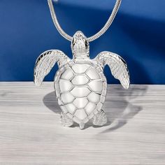 a silver turtle necklace with wings on a wooden table next to a blue wall and white background