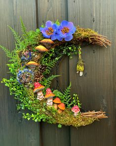 a wreath made out of moss with blue flowers and mushrooms on the front, hanging from a wooden fence