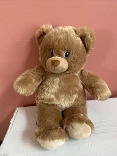 a brown teddy bear sitting on top of a white box with pink wall in the background