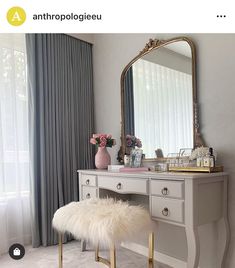 a vanity with a mirror, stool and flowers on it in front of a window