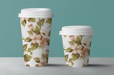 two coffee cups sitting next to each other on top of a gray surface with green leaves and flowers