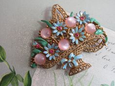 "Here's a vintage flower basket brooch featuring pink  moon glow cabochons and blue enamel flowers with pink rhinestone centers.  The setting is gold tone metal with a ribbon and bow enamel at the base.  It's in nice condition, and has a working safety clasp on the back of the basket.  Measures 2.1/4\" high and 2.3/4\" across.  Not signed, but most likely by CORO.  Please see the picture with a coin for size comparison, as items may appear larger than they actually are. This is necessary to prov Vintage Pink Collectible Brooches, Pink Costume Jewelry Brooch For Gift, Pink Costume Jewelry Brooches For Gifts, Pink Flower Enamel Brooch, Vintage Pink Enamel Brooches, Tahitian Pearl Pendant, Enamel Flowers, Flower Baskets, Tahitian Black Pearls