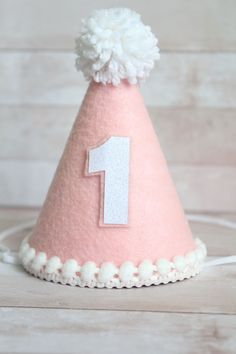 a pink felt party hat with a white number one on it and pom - poms around the top