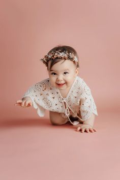 a smiling baby girl wearing a white dress and headband on her stomach, in front of a pink background