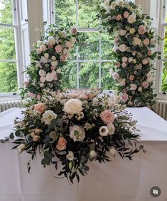 an arrangement of flowers sits on a table in front of a window with greenery