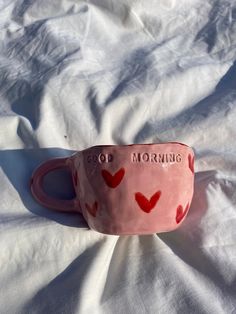 a pink coffee cup with red hearts on it sitting on a white sheet that says, good morning