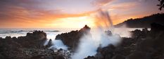 the sun is setting over an ocean with rocks and water spewing from it