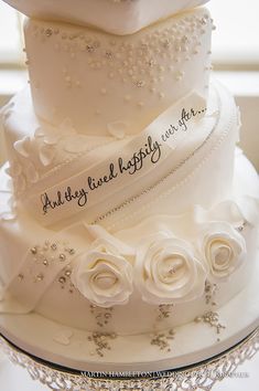 three tiered wedding cake with white roses and pearls on the bottom, decorated with an inscription