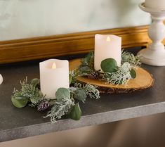 two white candles sitting on top of a table next to greenery and pine cones