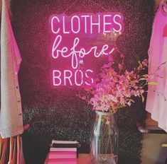 a pink neon sign that says clothes before bros on the wall next to a vase with flowers