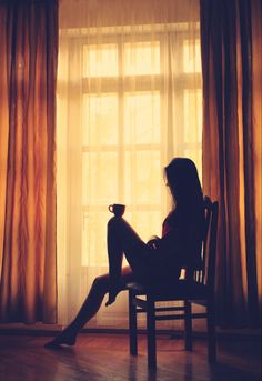 a woman sitting in a chair holding a coffee cup and looking out the window at the sun