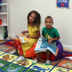 two children sitting on bean bag chairs in a playroom reading books and smiling at the camera