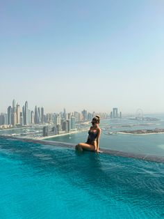 a woman sitting on the edge of a swimming pool in front of a cityscape