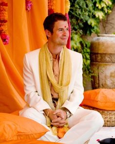 a man in a white suit and yellow shawl sitting on the ground next to orange drapes