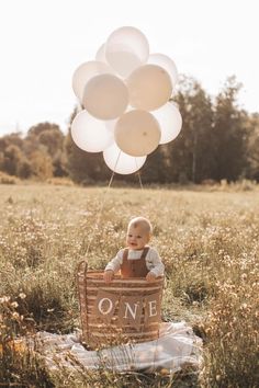 a baby sitting in a basket with white balloons floating above it and the words one written on it