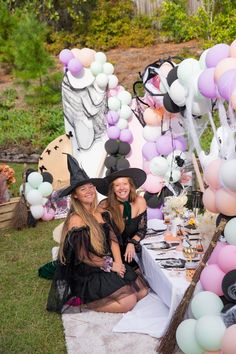 two women in witches costumes sitting at a table with balloons and decorations on the ground