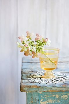 a small glass vase with flowers in it sitting on an old wooden table next to a white wall