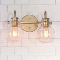 two light fixtures mounted on a white tiled wall in a bathroom with brass fixtures and clear glass globes