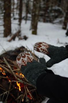 someone wearing mittens standing next to a fire in the woods with snow on it