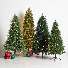 three artificial christmas trees with lights and presents on the floor in front of a white wall