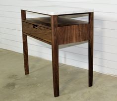 a wooden table with a white marble top and drawer on the bottom, in front of a white wall