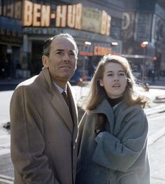 an older man and woman standing next to each other on the sidewalk in front of a building