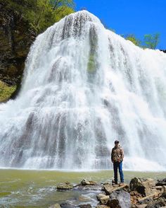 a man standing in front of a waterfall with his back to the camera and looking at it