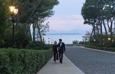 a man and woman walking down a path next to the ocean at dusk with lights on