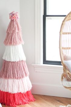 a pink, white and red christmas tree in front of a window with a wicker chair next to it