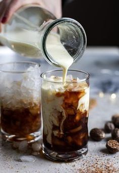 someone pouring milk into two glasses filled with ice and coffee beans on a counter top