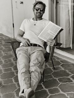 a man sitting in a chair reading a book and smiling at the camera while wearing sunglasses