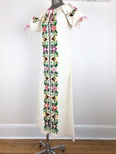 "1970s Mexican white cotton peasant dress has colorful floral embroidery down the front and around the sleeves, and a drawstring neckline and sleeve openings with pink braided yarn. Condition Overall very good, has some dye bleed on the front next to the purple flowers, a stain at the back right shoulder, and light grey discoloration at the hem from wear. All of the drawstring ends have fray. Measurements Bust 38\" Waist 35\" Hip 39\" Sleeve length 10\" Length 54\"" Spring Peasant Long Dress, Long Peasant Spring Dress, Spring Long Peasant Dress, Vintage Dresses With Multicolor Embroidery For Spring, Spring Folk Festival Embroidered Dress, Long Sleeve Folk Peasant Dress For Spring, Folk Style Cream Dress For Spring, Cream Folk Dress For Spring, Bohemian Cotton Embroidered Dress For Daywear