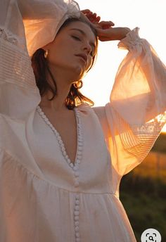 a woman wearing a white dress and holding her hands on her head with the sun shining behind her