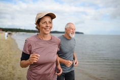 an older man and woman jogging on the beach together, with water in the background