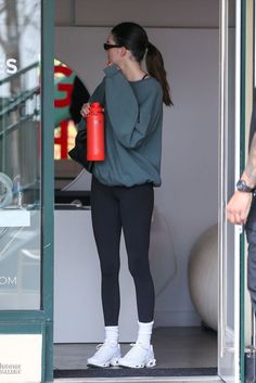 a woman in black leggings is holding a red water bottle and walking out of a building