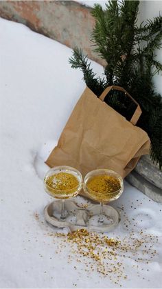 two wine glasses sitting in the snow next to a brown paper bag and pine tree